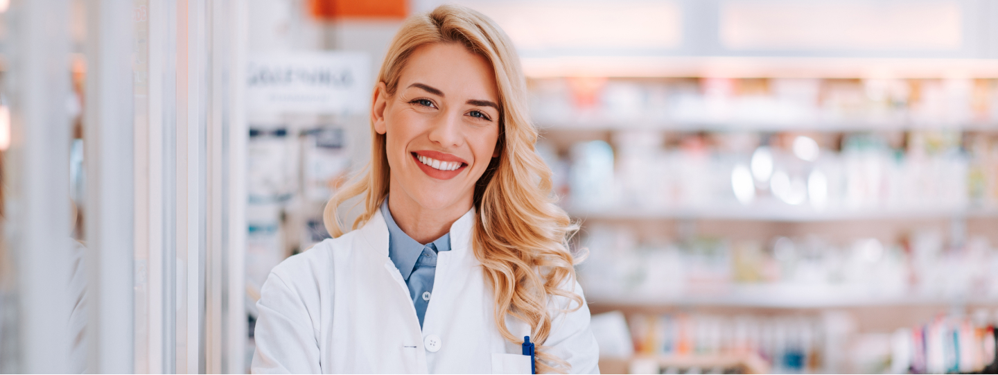 Eine lächelnde, blonde Frau im weißen Kittel steht mit verschränkten Armen vor einem Regal mit Medikamenten in einer Apotheke.